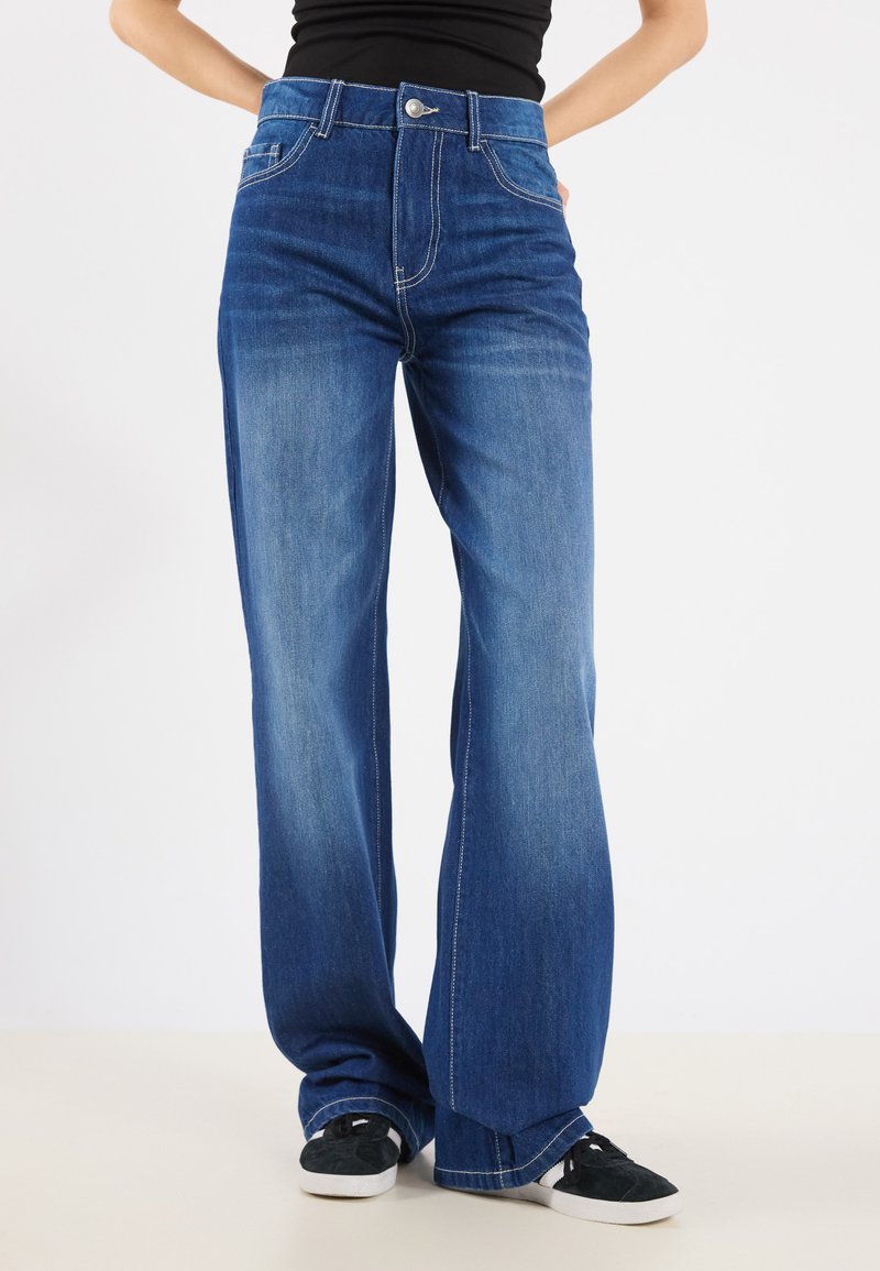 Jean en denim bleu à jambe large et taille haute, avec une fermeture à un bouton à l'avant, des coutures décoratives et une texture douce.