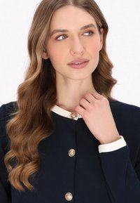 Navy jacket with a white collar, adorned with gold-tone buttons featuring decorative patterns. Smooth fabric and subtle texture details.