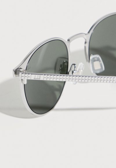 Γυαλιά ηλίου με ασημένιο μεταλλικό σκελετό, σκούρους φακούς και υφασμένο μπράτσο με το λογότυπο "dunhill" κοντά στο μεντεσέ.