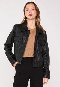 Veste en cuir noire avec col, fermeture par boutons et deux poches poitrine. Texture lisse et coupe ajustée, portée avec un haut marron.