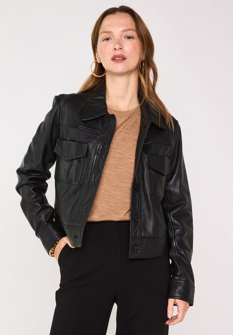 Veste en cuir noire avec col, fermeture par boutons et deux poches poitrine. Texture lisse et coupe ajustée, portée avec un haut marron.