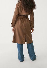 Bruine trenchcoat van gladde stof, met een ceintuur in de taille, een zijsplit en opgerolde manchetten, gecombineerd met wijde blauwe spijkerbroek