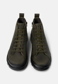 Bottines vert olive en cuir, avec des lacets, une fermeture éclair et une semelle en caoutchouc noir. Tige texturée avec un branding minimal.