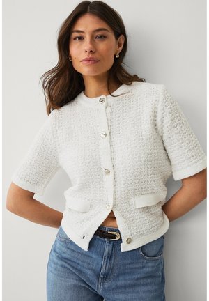 Vrouw draagt een wit gestructureerd kort mouwen vestje met gouden knopen en blauwe jeans, staand tegen een effen lichte muur.