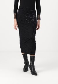 Jupe midi noire à motif floral, en tissu texturé. Portée avec un haut noir moulant à manches longues et des bottes en cuir noires montant jusqu'aux genoux.