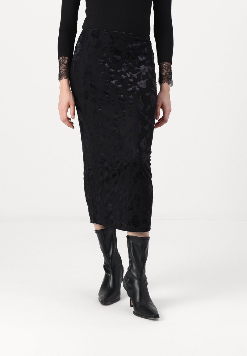 Jupe midi noire à motif floral, en tissu texturé. Portée avec un haut noir moulant à manches longues et des bottes en cuir noires montant jusqu'aux genoux.