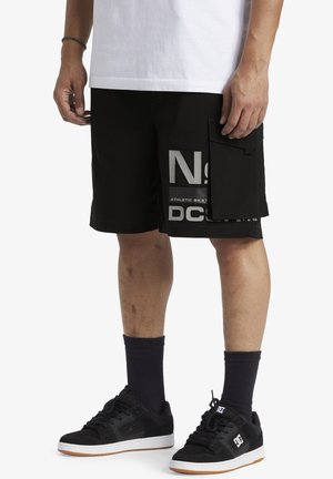 Schwarze Cargo-Shorts mit auffälligem Grafikdruck, weiten Taschen und einem strukturierten Stoff. Kombiniert mit schwarzen Sportschuhen und dunklen Socken.