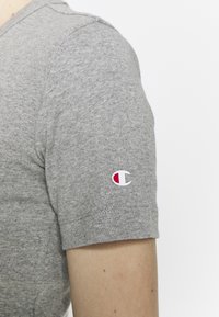 Šedé tričko s krátkým rukávem s žebrovanou texturou, které má malé bílo-červené logo na levém rukávu.