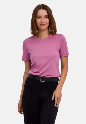 Vrouw met schouderlang bruin haar, gekleed in een roze shirt met korte mouwen en een zwarte broek met een zwarte riem, staand voor een witte achtergrond.
