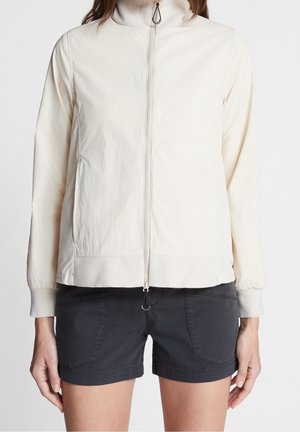 Kobieta ubrana w jasnobeżową kurtkę na zamek z wysokim kołnierzem oraz czarne szorty z przednimi kieszeniami na jednolitym tle.