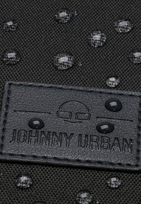 Svart texturerad tyg med vattendroppar, med en upphöjd svart läderetikett präglad med "JOHNNY URBAN" och grafiska detaljer.