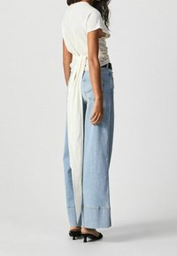 Biała koszula z krótkim rękawem z detalem w postaci wiązania z tyłu, zestawiona z luźnymi, jasnoniebieskimi dżinsami i czarnymi sandałami na obcasie.