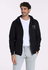 Haut à zip noir avec une capuche à cordon de serrage, arborant un petit logo sur la poitrine gauche, associé à un pantalon de survêtement gris clair.