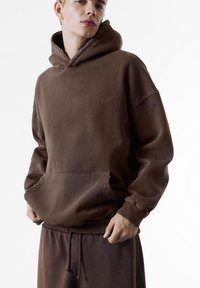 Bruine hoodie met een kangoeroezak, geribbelde manchetten en een trekkoord. Gemaakt van zachte stof, met een klein logo op de borst.