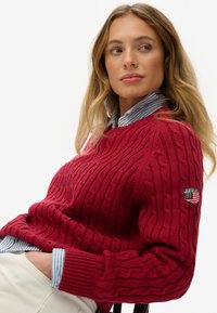 Czerwony sweter z grubym splotem, okrągłym dekoltem i ściągaczami na rękawach, ozdobiony naszywką z logo na lewym rękawie, noszony na paskowanej koszuli.