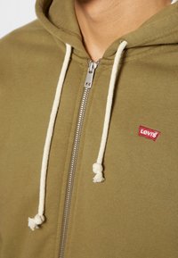 Felpa con zip color verde oliva, con lacci crema e un'etichetta rossa di Levi's sul davanti. Realizzata in un tessuto morbido e texturizzato.