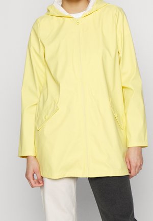 Personne portant un imperméable à capuche jaune clair avec fermeture éclair frontale et poches à rabat, assorti à un pantalon bicolore, sur un fond uni.