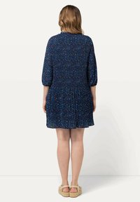 Kvinna som står med ryggen mot kameran, iförd en marinblå klänning med små flerfärgade blommor och beige plattformssandaletter.