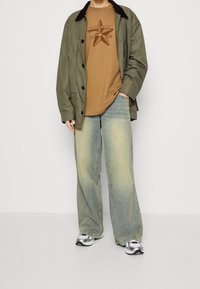 Oliivinvihreä takki, ruskea graafinen t-paita, haalistuneet siniset leveälahkeiset farkut ja hopeiset lenkkarit. Teksturoidut materiaalit selkeillä värit kontrasteilla.