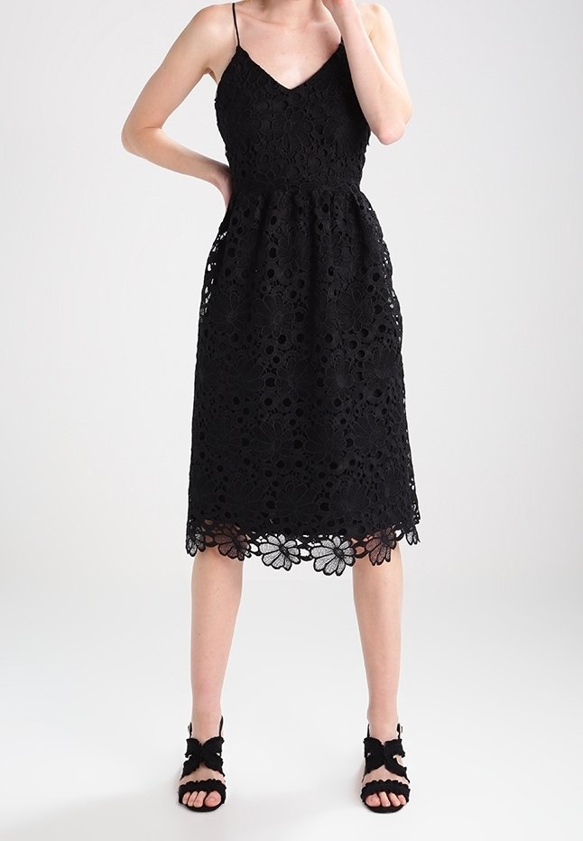 Femme portant une robe en dentelle noire mi-longue avec de fines bretelles et des motifs floraux, assortie à des talons ouverts noirs avec des nœuds.