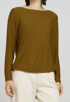 Kobieta ubrana w brązową bluzkę z długim rękawem i dekoltem w łódkę oraz kremowe spodnie na jednolitym tle.