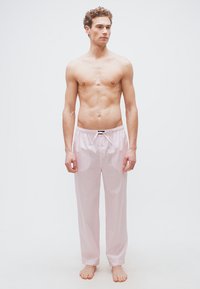 Νέος άνδρας όρθιος, ξυπόλητος, χωρίς πουκάμισο, φοράει ανοιχτό ροζ και λευκή ριγωτή φαρδιά πυτζάμα, μπροστά σε απλό λευκό φόντο.