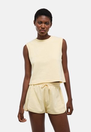 Femme portant un top sans manches jaune clair et un short assorti à cordon, debout devant un fond clair uni.
