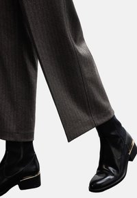 Osoba nosząca brązowe spodnie w rybi wzór i czarne skórzane botki za kostkę z niskim obcasem i metalicznym akcentem na podeszwie.