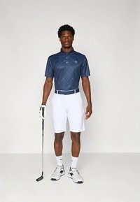 Námornícka tmavomodrá golfová polokošeľa s krátkym rukávom, s diamantovým vzorom a emblémom, v kombinácii s bielymi šortkami. Obsahuje biely opasok a golfovú palicu. Textúrované topánky.