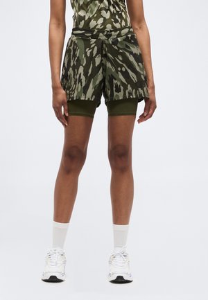 Persoană purtând pantaloni scurți suprapuşi cu model verde şi negru, încălţată cu pantofi sport albi și şosete albe până la jumătatea gambei, stând pe un fundal simplu.