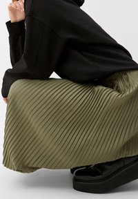 Falda plisada verde oliva de textura fluida, combinada con una sudadera negra de gran tamaño y zapatos de plataforma negros. Detallada con pliegues verticales.