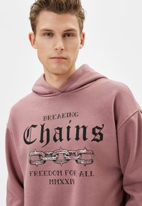 Rosa sweatshirt med huva, prydd med svart text "BREAKING Chains FREEDOM FOR ALL MMXXIV" och en grafik av en kedja. Mjuk, slät textur.