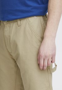 Beige bomull cargo bukser med sidelommer, synlige sømmer og et lite logoetikett på midjen, sammen med en blå kortermet t-skjorte.