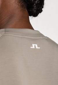 Világostaupe színű póló kerek nyakkivágással, a hátán fehér logóval. Az anyaga sima megjelenésű, finom textúrával.