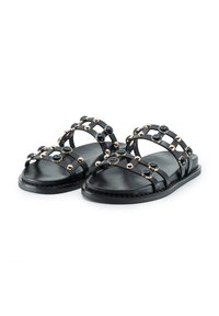 Schwarze flache Sandalen aus Leder mit zwei überkreuzten Riemen, die mit goldenen und schwarzen dekorativen Nieten verziert sind. Glatte Textur und minimales Design.