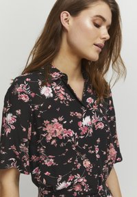 Blouse noire à motifs floraux, avec des fleurs roses et blanches, des manches courtes, un col et des boutons sur le devant. Fabriquée dans un tissu lisse.