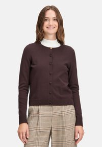 Dark brown cardigan with a round neckline, buttons down the front, ribbed cuffs and hem, worn over a white high-neck top and patterned plaid trousers.