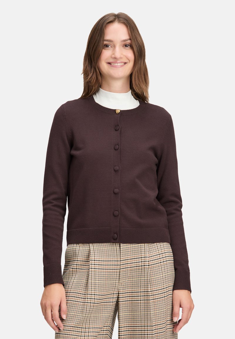Dark brown cardigan with a round neckline, buttons down the front, ribbed cuffs and hem, worn over a white high-neck top and patterned plaid trousers.