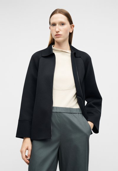 Νεαρή γυναίκα με ίσια μαλλιά που φοράει μαύρο μπουφάν με φερμουάρ, κρεμ μπλουζάκι και χαλαρό γκρι παντελόνι, στέκεται με το ένα χέρι στην τσέπη.