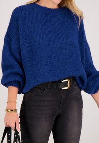 Vrouw draagt een blauwe grofgebreide trui met ballonmouwen, zwarte riem met gouden gesp, zwarte spijkerbroek, gouden armbanden en houdt een zwarte gestikte tas vast.