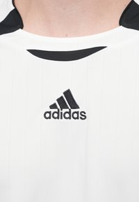Biała koszulka sportowa z czarnymi akcentami, wykonana z teksturowanej tkaniny. Posiada pionowe paski oraz wyhaftowane czarne logo Adidas na piersi.