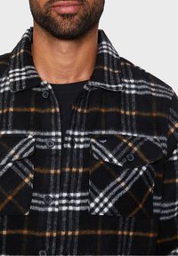 Hombre vistiendo una camisa de cuadros en negro, blanco y marrón sobre una camiseta negra, mostrando el cuello y la parte superior del pecho.