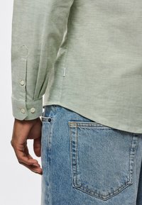 Ανοιχτοπράσινο πουκάμισο με μακριά μανίκια και κουμπωτά μανσέτα, συνδυασμένο με μπ λε τζιν τζιν. Το πουκάμισο διαθέτει ίσια τελείωμα και διακριτή υφή.