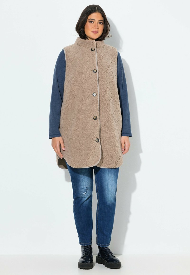 Gilet imbottito beige con alto colletto, bottoni frontali e tasche laterali, indossato sopra una camicia a maniche lunghe blu. Abbinato a jeans blu e stivali.