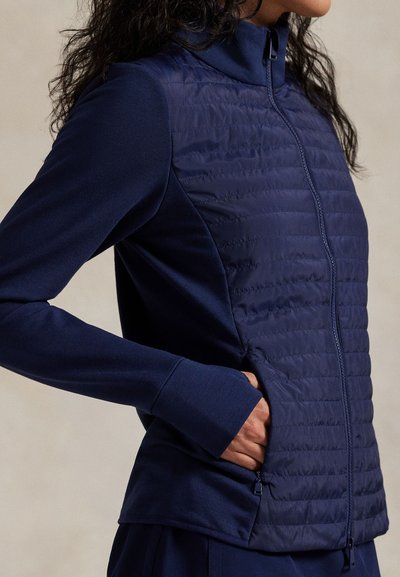 Μπλε ναυτικό μπουφάν με φερμουάρ μπροστά, που διαθέτει επάνω τμήμα με καπιτονέ και λείες κάτω μανσέτες. Εφαρμοστή γραμμή με πλαϊνές τσέπες με φερμουάρ.