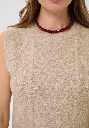 Femme portant un pull sans manches beige en maille torsadée et un collier de perles rouge foncé, avec de longs cheveux châtain clair visibles.