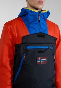 Chaqueta colorida con capucha azul, mangas naranjas y torso negro con acentos azules. Incluye múltiples bolsillos con cremallera y un parche de la bandera de Noruega.
