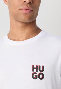 Bela bombažna majica s kratkimi rokavi, ki ima okroglo izrez in črno-rdeč grafični tisk besede "HUGO" na levem prsnem delu. Gladka tekstura.
