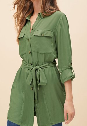 Chemise verte à boutons avec deux poches poitrine, taille nouée, manches retroussables et texture lisse. Portée avec un jean en denim.