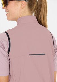 Hellrosa Sportweste mit hohem Kragen, die schwarze Akzente und einen reflektierenden Streifen auf der Rückseite aufweist. Glatte Textur und reguläre Passform.